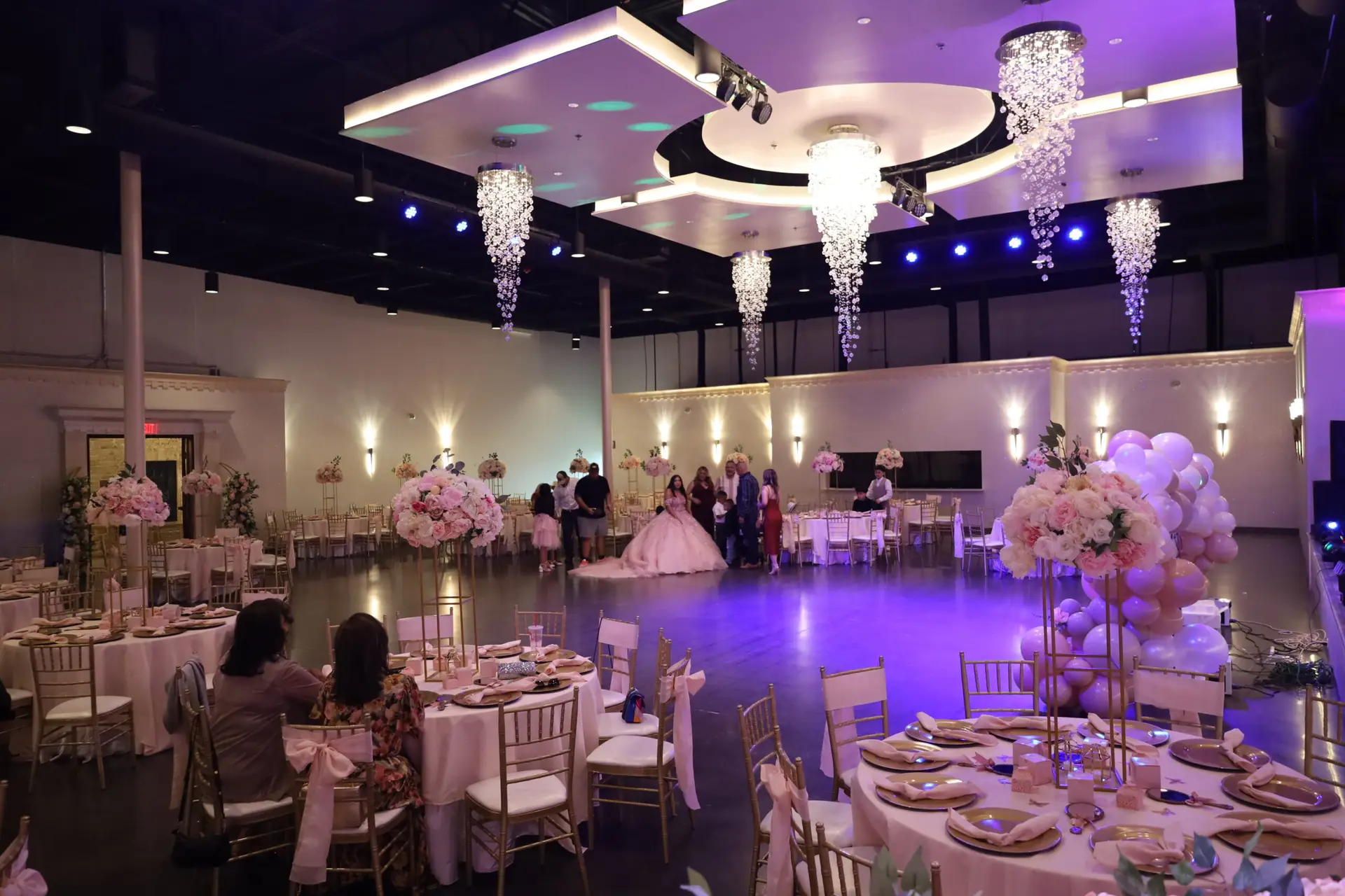 Quinceañera party in a majestic banquet hall with pink and gold decor, featuring elegant table settings, floral arrangements, and ambient lighting