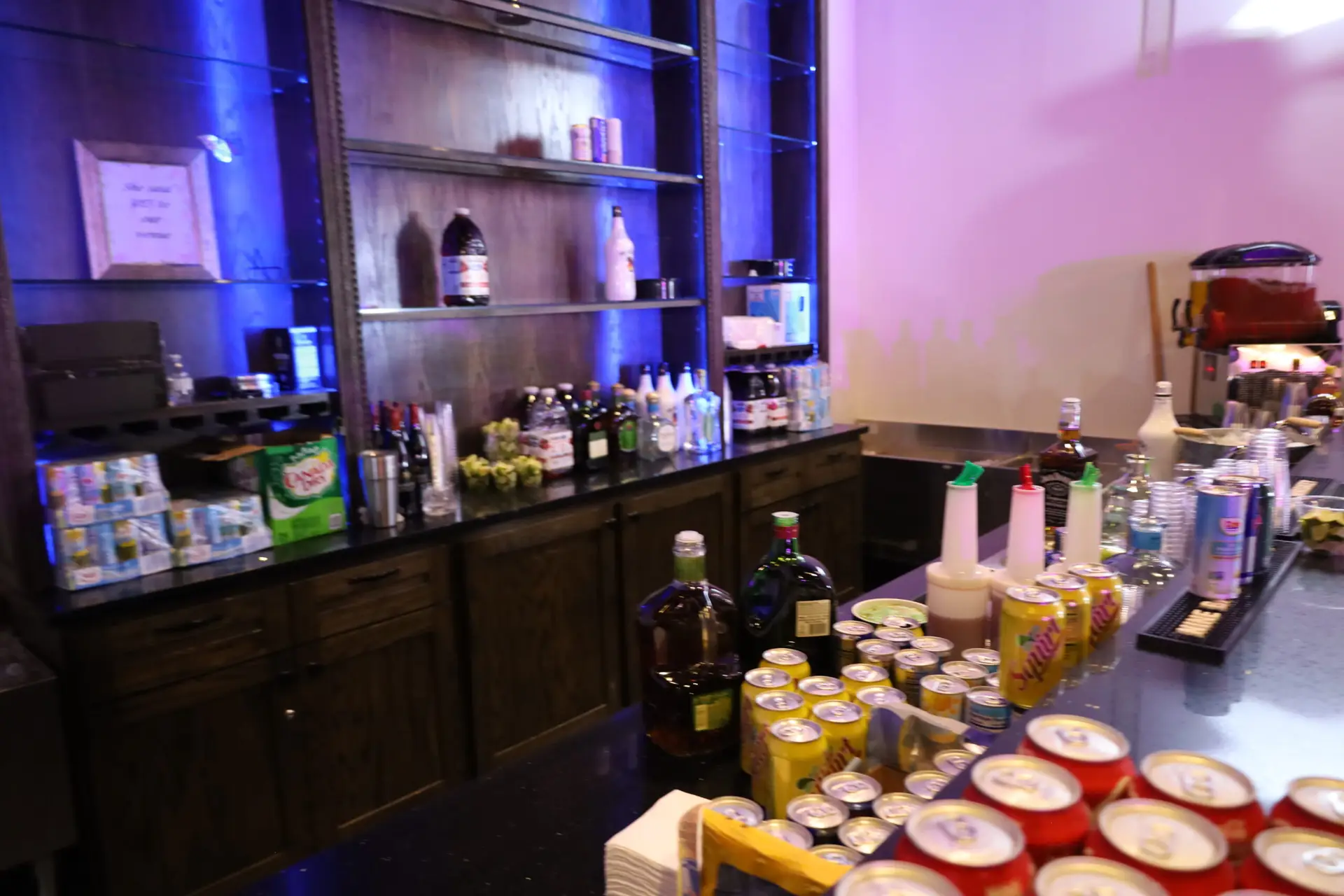 Large wet bar with sleek black granite countertops, vibrant LED underlighting, stylish tiled backsplash, built-in coolers, beer troughs, glass shelves illuminated with LED backlighting, and an ice machine for events and gatherings